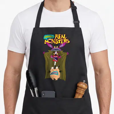 Aaahh boy monster apron - Three cartoon monsters stacked vertically under a green cloak below yellow Real Monsters text — shown on a black cotton canvas apron against a white background