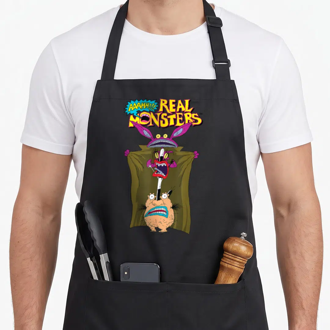 Aaahh boy monster apron - Three cartoon monsters stacked vertically under a green cloak below yellow Real Monsters text — shown on a black cotton canvas apron against a white background
