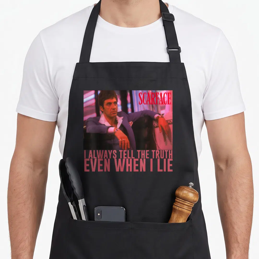 Scarface scaredy apron - Tony Montana photo with SCARFACE logo and truth lie quote text — shown on a black cotton canvas apron against a white background