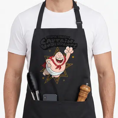 Underpant captain apron - Arched text above a peach cartoon man in white underwear and red cape flying in front — shown on a black cotton canvas apron against a white background