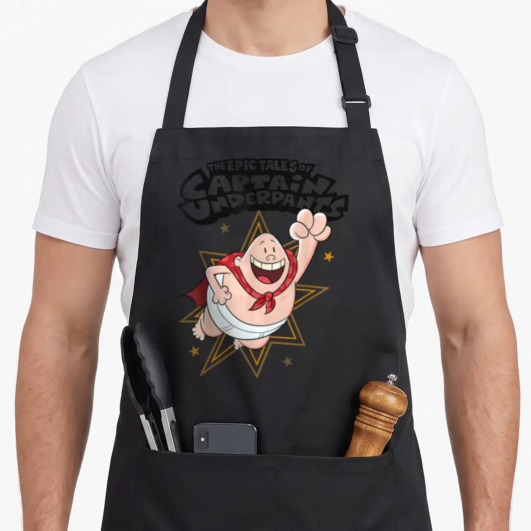 Underpant captain apron - Arched text above a peach cartoon man in white underwear and red cape flying in front — shown on a black cotton canvas apron against a white background