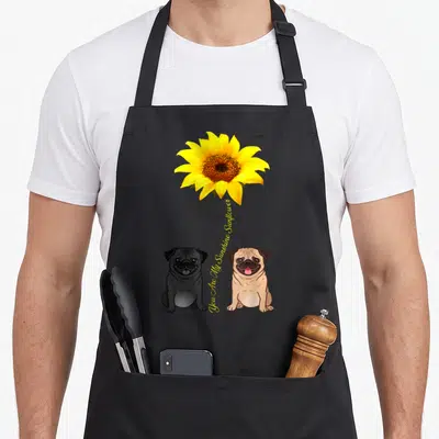 Pug sunflower apron - Two cartoon pugs sitting below a yellow sunflower with vertical text You Are My Sunshine Sunflower — shown on a black cotton canvas apron against a white background
