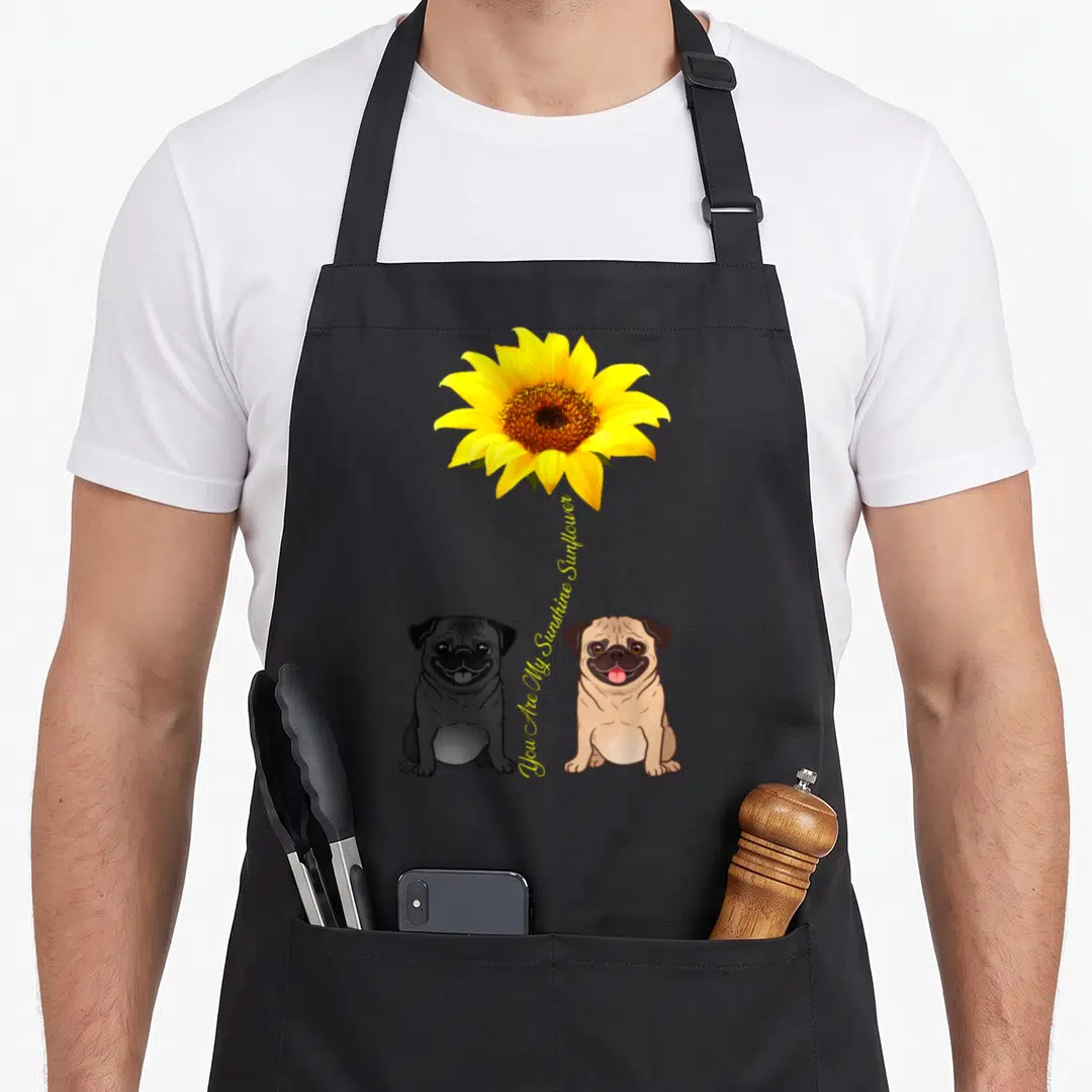 Pug sunflower apron - Two cartoon pugs sitting below a yellow sunflower with vertical text You Are My Sunshine Sunflower — shown on a black cotton canvas apron against a white background