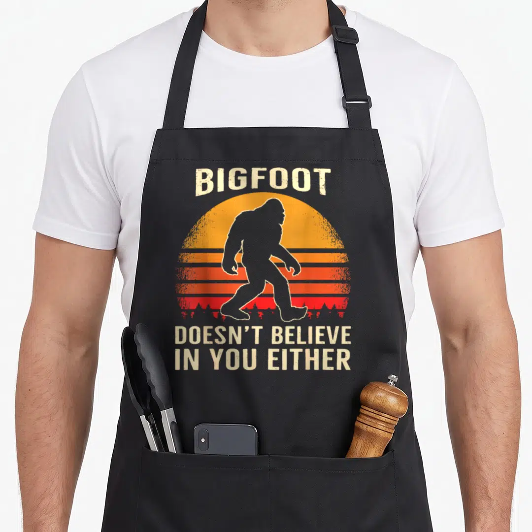 Either sasquatch bigfoot apron - Retro Bigfoot silhouette against orange and red striped sun with sarcastic text — shown on a black cotton canvas apron against a white background