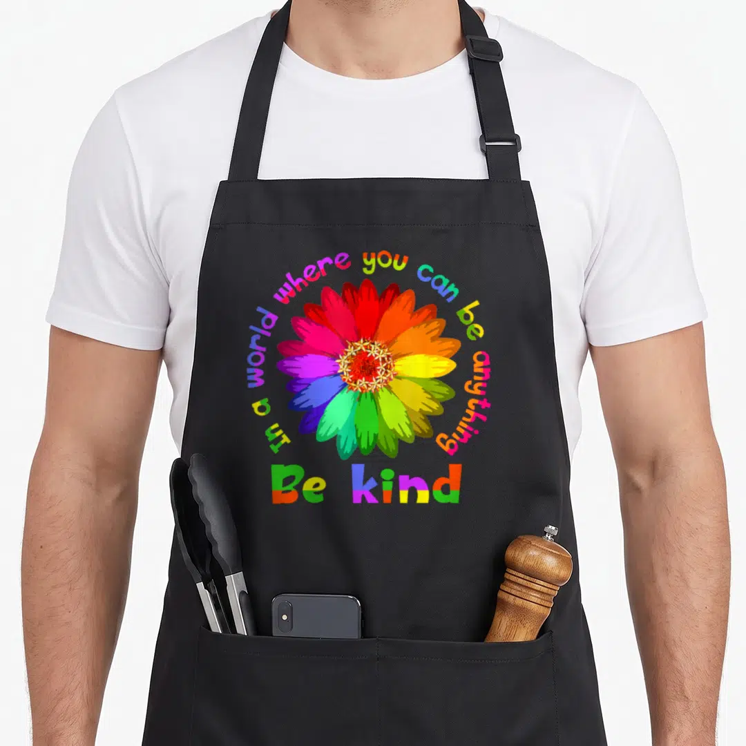 Anything unity apron - Rainbow cartoon flower with arched text In a world where you can be anything and block text Be — shown on a black cotton canvas apron against a white background
