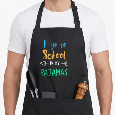 School pajamas novelty apron - Stacked text reading I go to school in my pajamas with a white arrow in blue orange — shown on a black cotton canvas apron against a white background