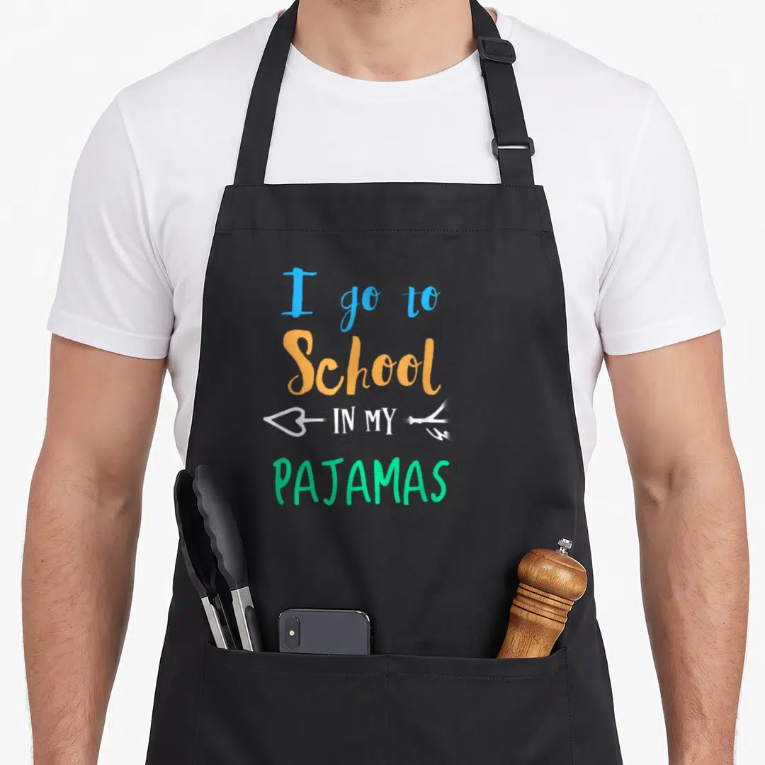 School pajamas novelty apron - Stacked text reading I go to school in my pajamas with a white arrow in blue orange — shown on a black cotton canvas apron against a white background