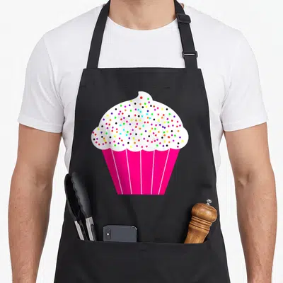 Food cupcake apron - Cartoon cupcake with white frosting, multicolored sprinkles, and pink pleated liner — shown on a black cotton canvas apron against a white background
