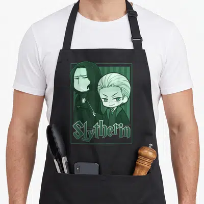 Chibi potter apron - Two men in green monochrome standing before vertical stripes with Slytherin text below — shown on a black cotton canvas apron against a white background