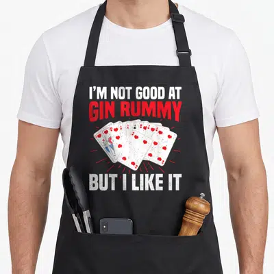 Gin rummy game aprons - Distressed text reading I'M NOT GOOD AT GIN RUMMY BUT I LIKE IT above and below a fan of hearts — shown on a black cotton canvas apron against a white background
