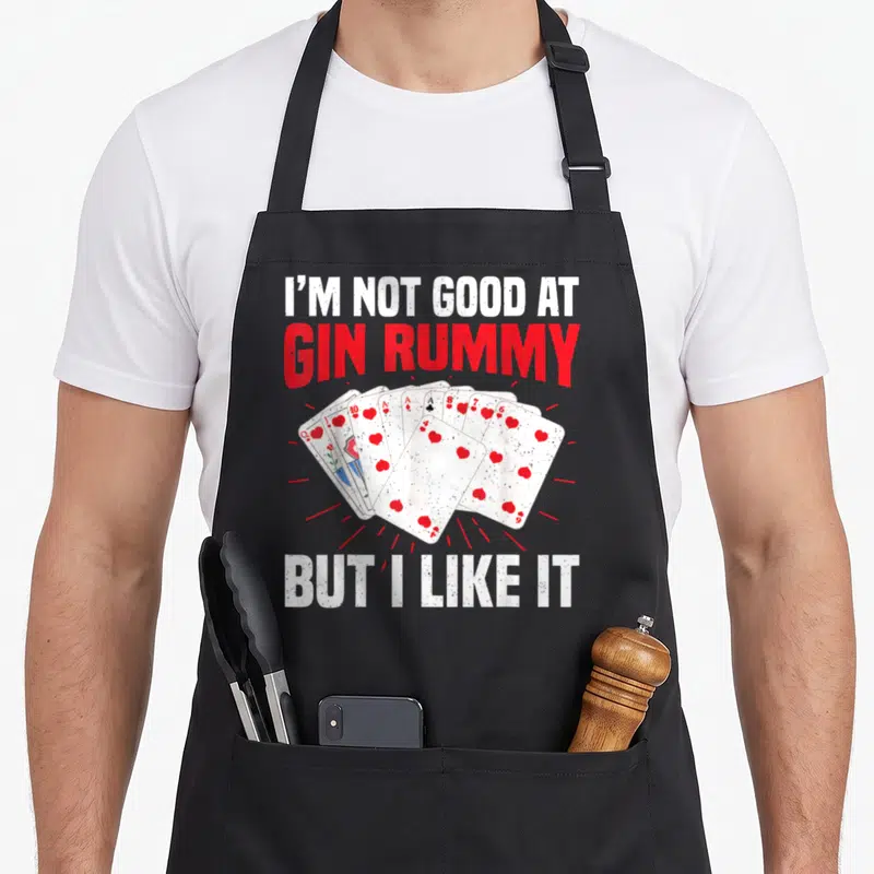 Gin rummy game aprons - Distressed text reading I'M NOT GOOD AT GIN RUMMY BUT I LIKE IT above and below a fan of hearts — shown on a black cotton canvas apron against a white background