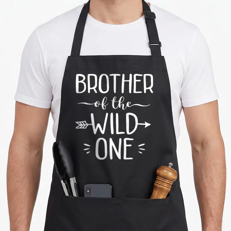 Brother wild birthday apron - White stacked text reading Brother of the Wild One with an arrow through Wild — shown on a black cotton canvas apron against a white background