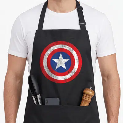 Funny shield captain apron - Distressed circular shield with red white rings and blue center containing a white star — shown on a black cotton canvas apron against a white background