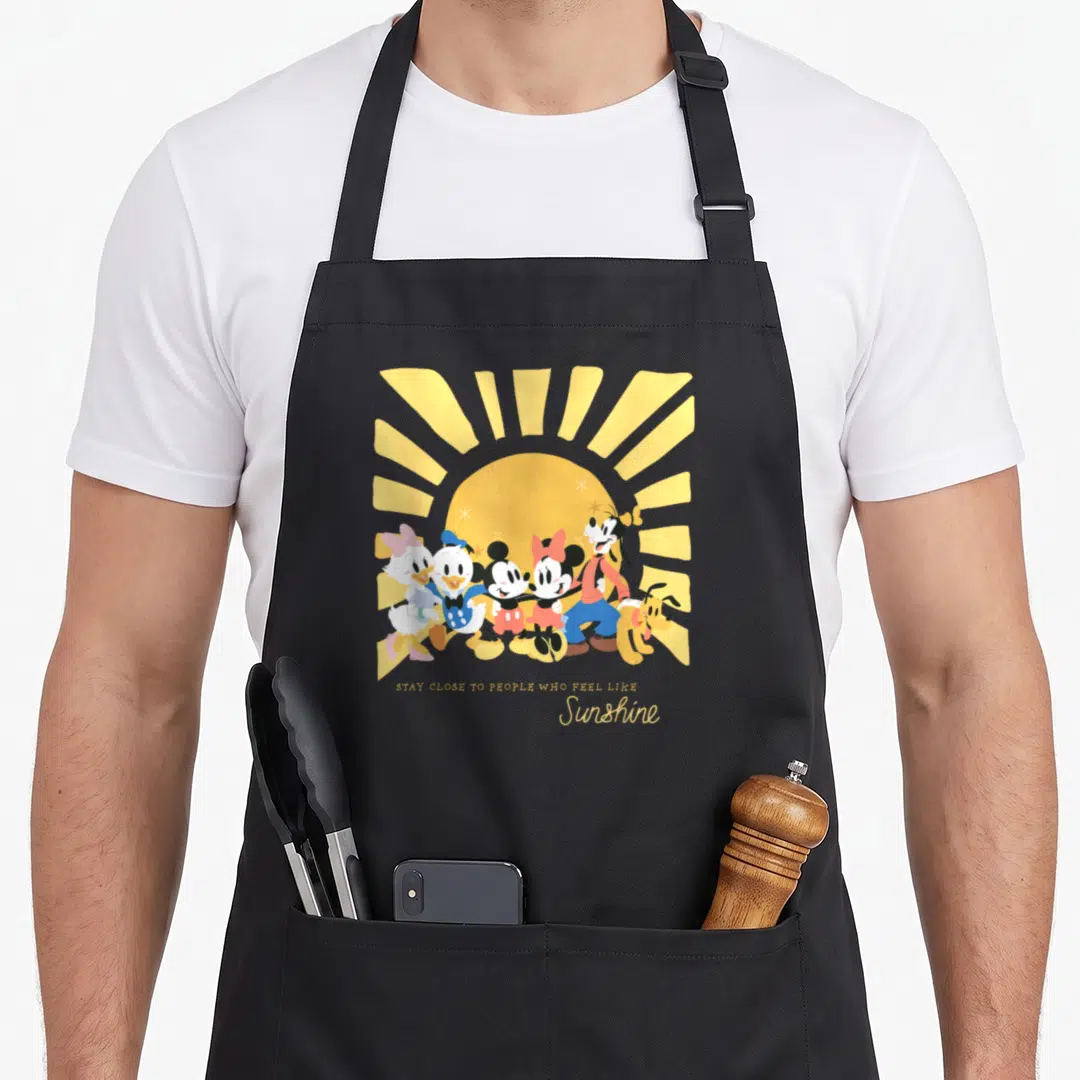 Feel friends apron - Mickey and Friends group under yellow sun with Stay Close To People Who Feel Like Sunshine text — shown on a black cotton canvas apron against a white background