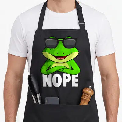 Critter frog aprons - Green cartoon frog with black sunglasses and white NOPE text — shown on a black cotton canvas apron against a white background