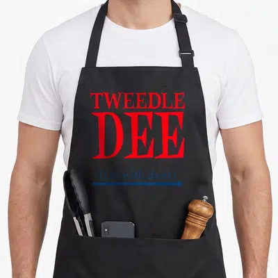 Dee funny halloween aprons - Red Tweedle Dee text with blue i'm with dum caption and right-pointing arrow — shown on a black cotton canvas apron against a white background