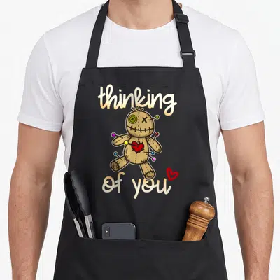Thinking doll apron - Brown voodoo doll with pins and red heart between thinking of you script text — shown on a black cotton canvas apron against a white background