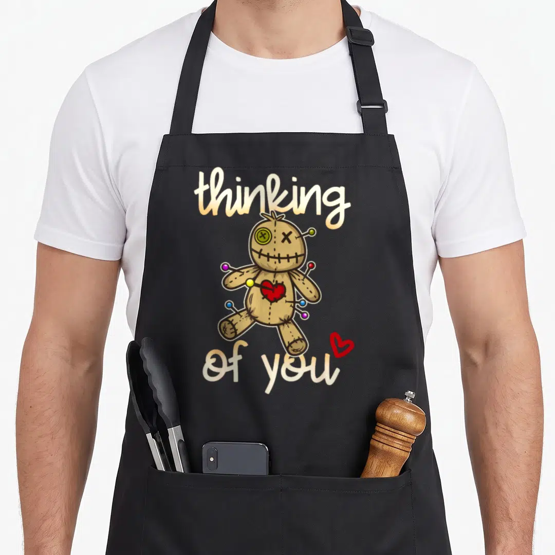 Thinking doll apron - Brown voodoo doll with pins and red heart between thinking of you script text — shown on a black cotton canvas apron against a white background