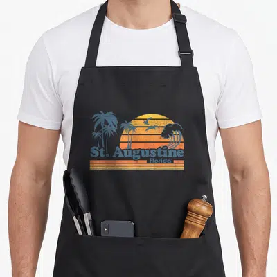Augustine spring aprons - Retro orange and blue sunset with palm trees and St Augustine Florida text — shown on a black cotton canvas apron against a white background