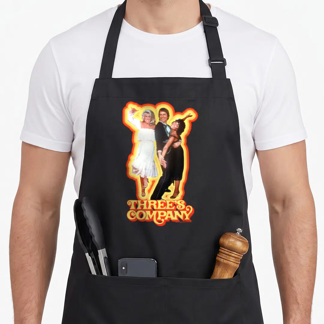 Dancing 80s apron - Three people in photo style inside yellow orange border with stacked THREE'S COMPANY text below — shown on a black cotton canvas apron against a white background