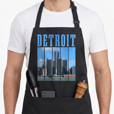 Detroit skyline pride apron - DETOIT text above five vertical panels showing city skyline in blue grey and brown colors — shown on a black cotton canvas apron against a white background
