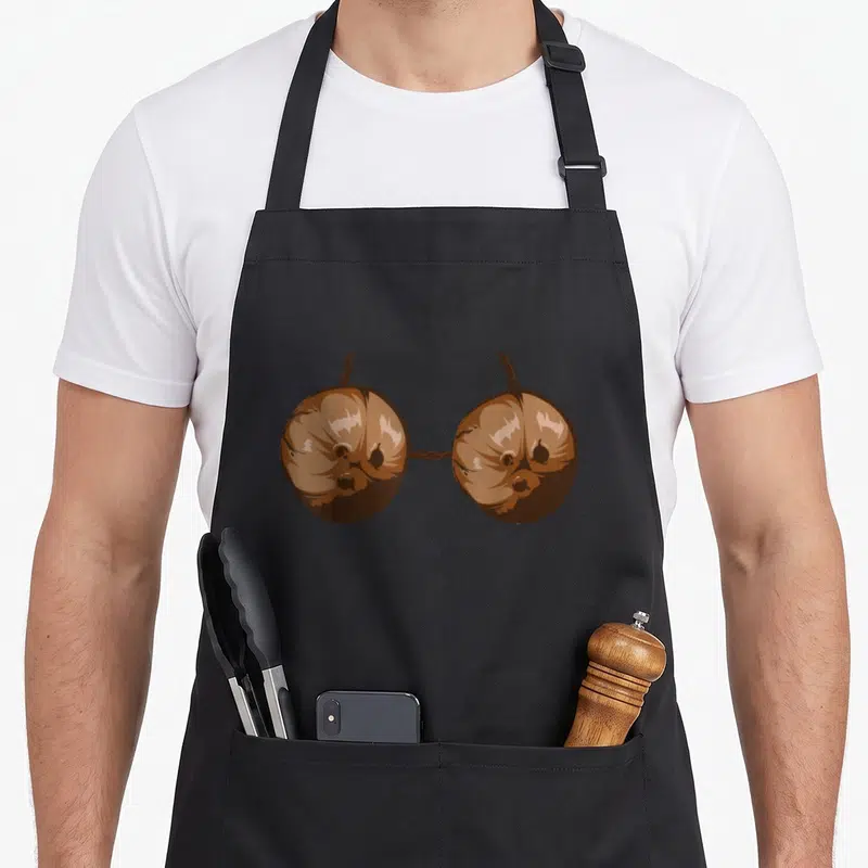 Summer halloween apron - Two brown cartoon coconuts connected by string forming a bra shape on black fabric — shown on a black cotton canvas apron against a white background