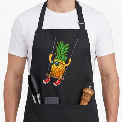 Swinging pineapple apron - Cartoon pineapple on swing with sunglasses and red shoes — shown on a black cotton canvas apron against a white background