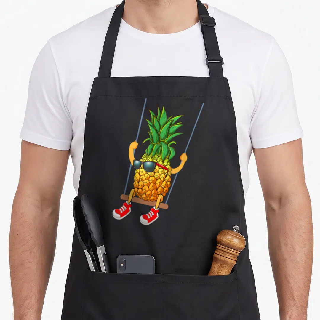 Swinging pineapple apron - Cartoon pineapple on swing with sunglasses and red shoes — shown on a black cotton canvas apron against a white background