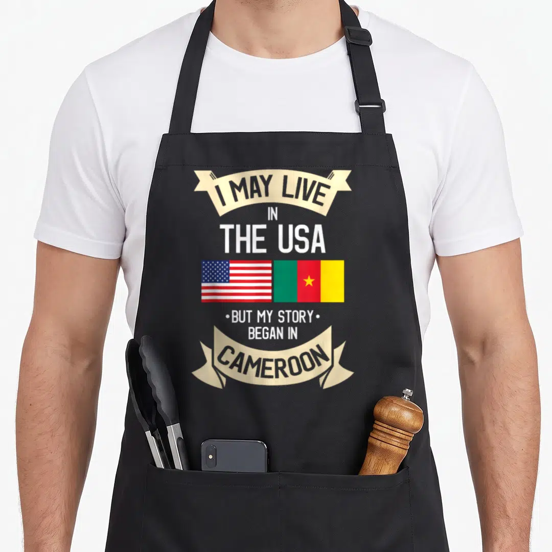 Cameroon american flag apron - I May Live In The USA But My Story Began In Cameroon text with US and Cameroon flags — shown on a black cotton canvas apron against a white background