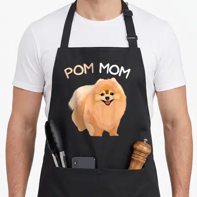 Pomeranian mama apron - Arched POM MOM text above a smiling orange cartoon Pomeranian dog — shown on a black cotton canvas apron against a white background