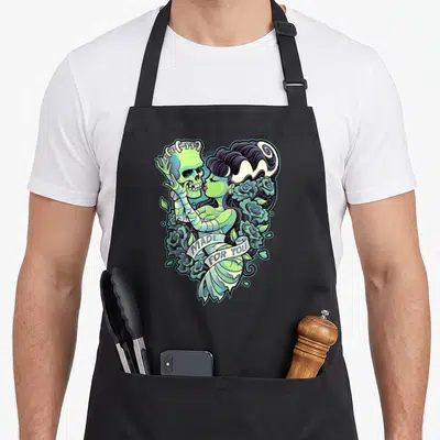 Frankenstein bride apron - Cartoon skeleton and woman in green and purple forming a heart with roses and text — shown on a black cotton canvas apron against a white background