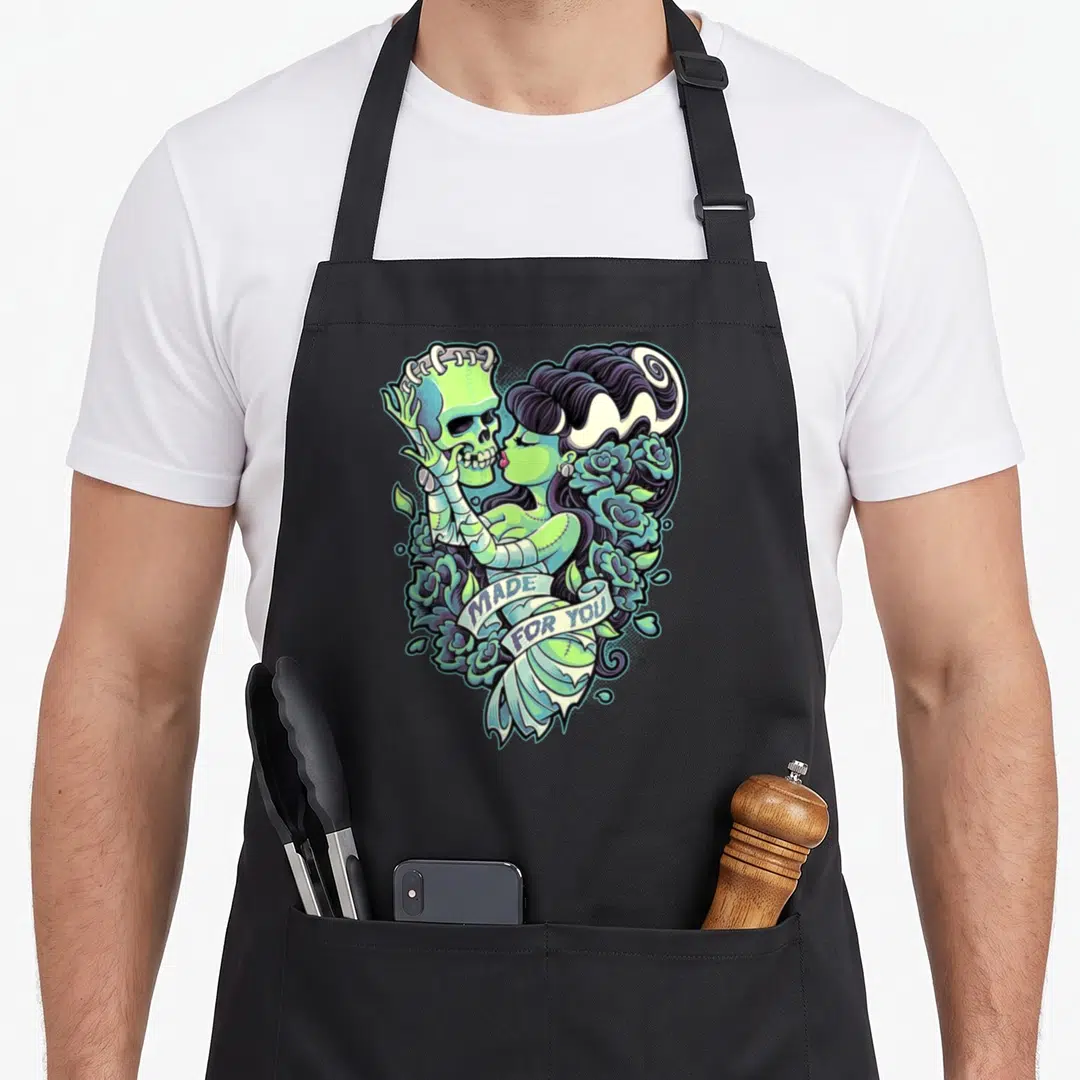 Frankenstein bride apron - Cartoon skeleton and woman in green and purple forming a heart with roses and text — shown on a black cotton canvas apron against a white background