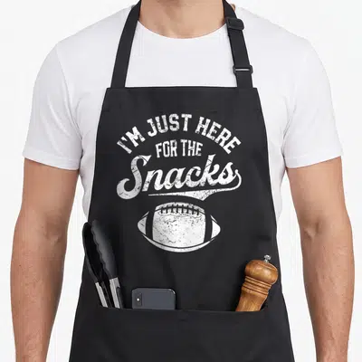 Snack football apron - White distressed text I'M JUST HERE FOR THE Snacks above a football graphic — shown on a black cotton canvas apron against a white background