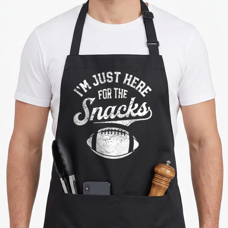 Snack football apron - White distressed text I'M JUST HERE FOR THE Snacks above a football graphic — shown on a black cotton canvas apron against a white background