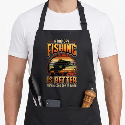 Better fisher fishing aprons - Retro sun graphic with black fish silhouette and orange gradient text reading A BAD DAY — shown on a black cotton canvas apron against a white background