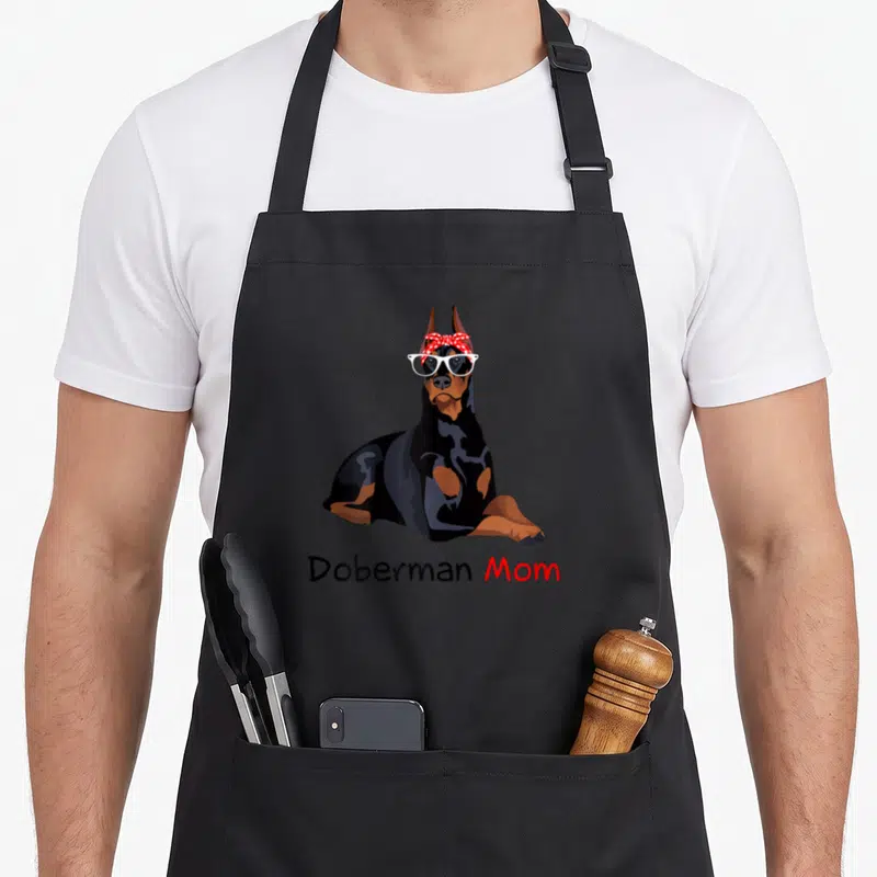 Doberman bandana dog apron - Cartoon Doberman with white glasses and red bow wearing Doberman Mom text — shown on a black cotton canvas apron against a white background
