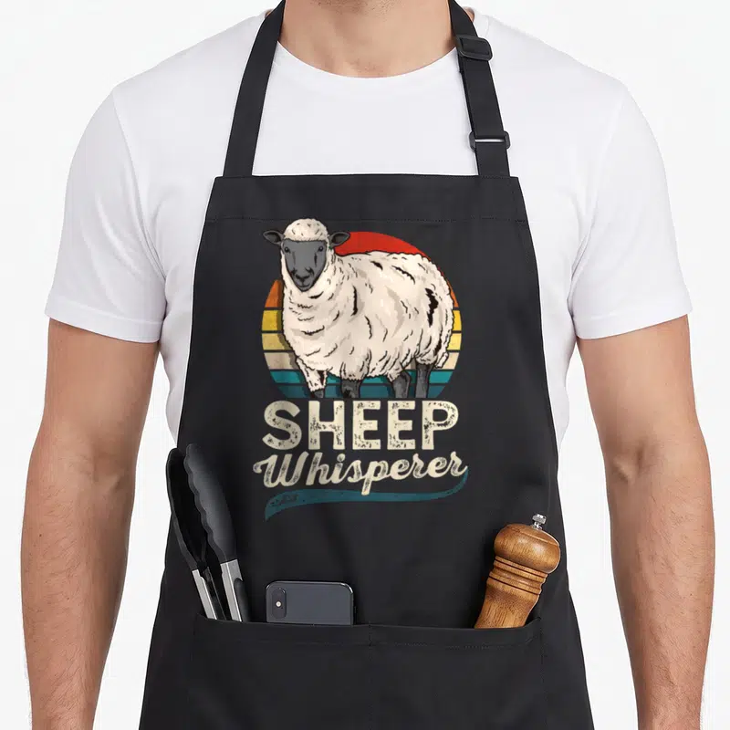 Sheep farmer apron - Retro sheep illustration over red orange yellow teal striped circle with SHEEP Whisperer text below — shown on a black cotton canvas apron against a white background