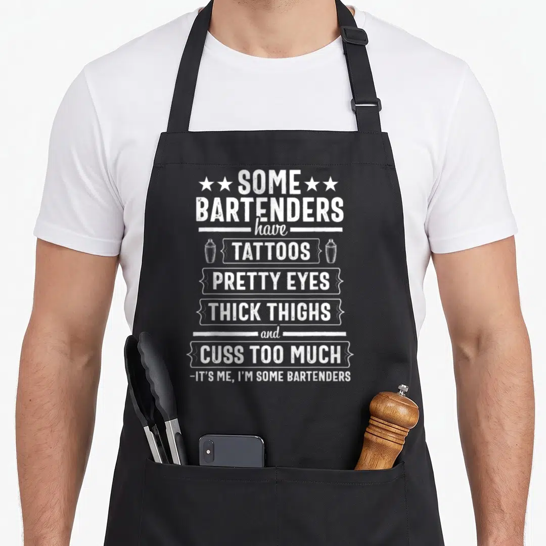 Thigh bartender apron - White stacked text listing bartender traits with two cocktail shakers flanking the center word — shown on a black cotton canvas apron against a white background