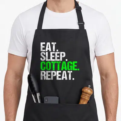 Eat cottage apron - Stacked EAT SLEEP COTTAGE REPEAT text in white and green distressed font — shown on a black cotton canvas apron against a white background