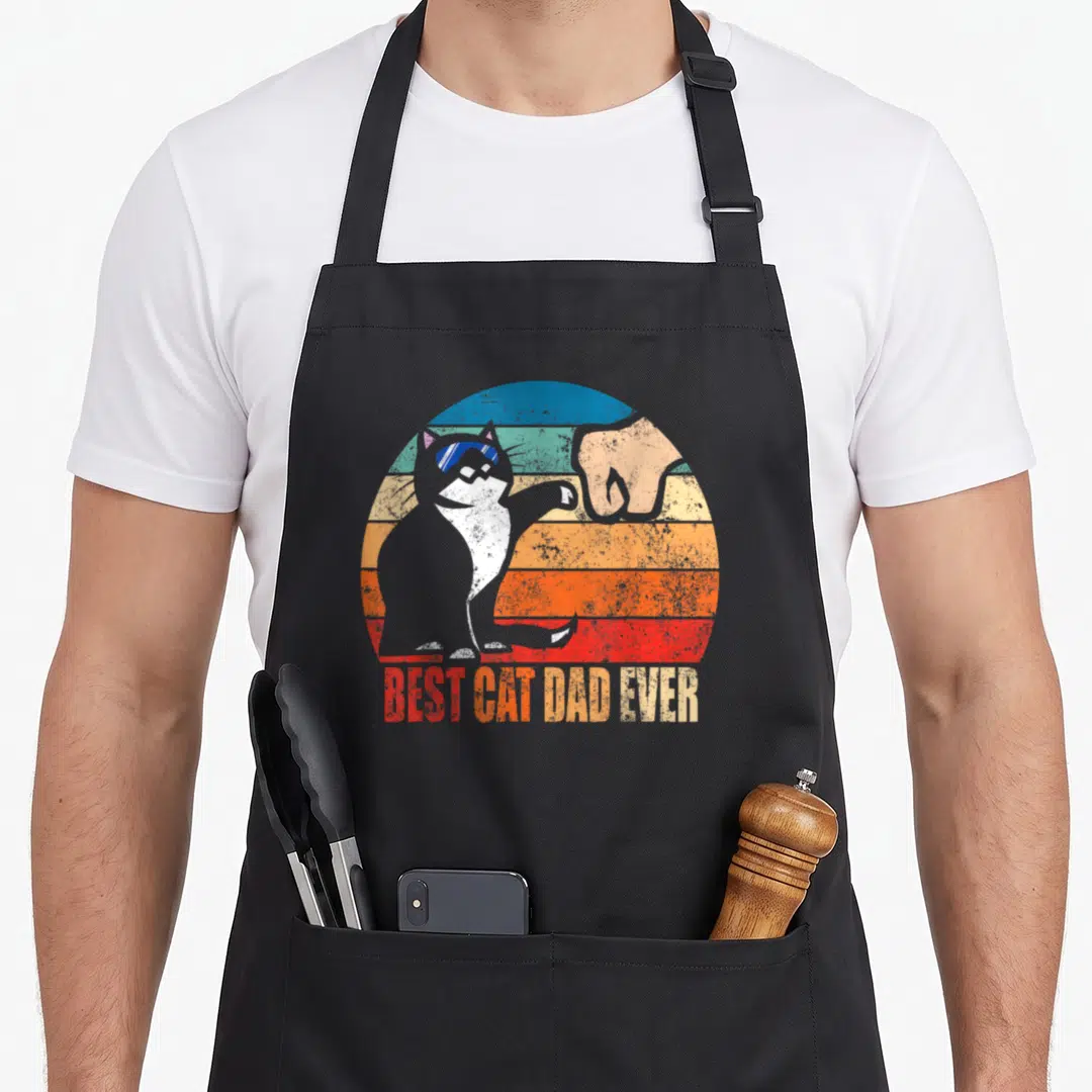 Cat father day apron - Grey cat fist bumping human hand in striped circle with BEST CAT DAD EVER text — shown on a black cotton canvas apron against a white background