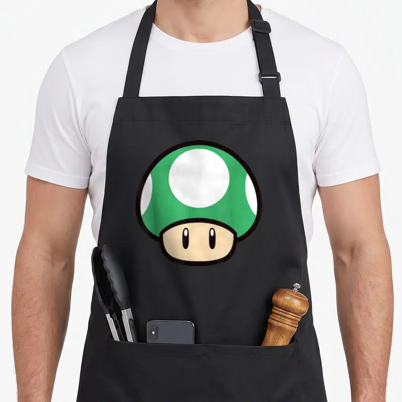 Funny mushroom apron - Green cartoon mushroom with white spots and beige stem — shown on a black cotton canvas apron against a white background