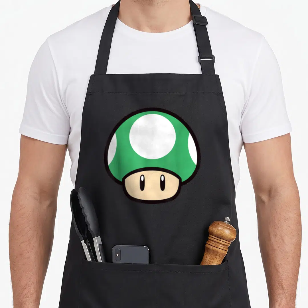 Funny mushroom apron - Green cartoon mushroom with white spots and beige stem — shown on a black cotton canvas apron against a white background