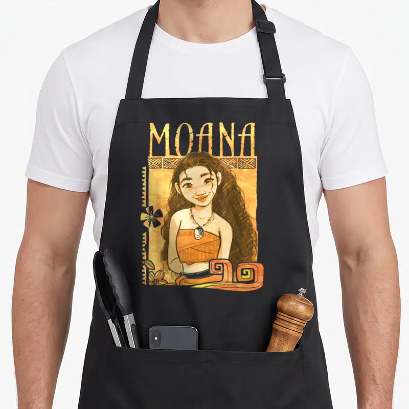 Artistic kitchen island apron - Cartoon Moana portrait with tribal border, orange background, and flower accent — shown on a black cotton canvas apron against a white background