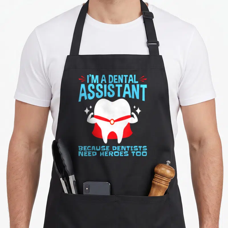 Dental funny comfort apron - Superhero tooth flexing muscles with red cape and blue text about dental assistants — shown on a black cotton canvas apron against a white background