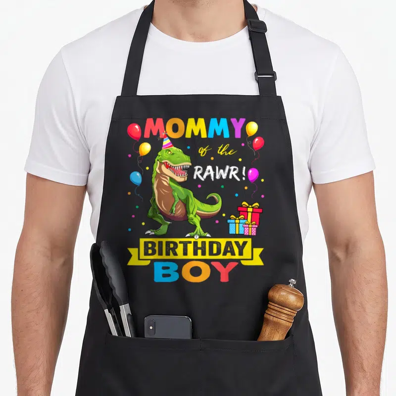 Birthday raptor dinosaur apron - Green T-Rex wearing party hat with MOMMY of the RAWR! BIRTHDAY BOY text and balloons — shown on a black cotton canvas apron against a white background