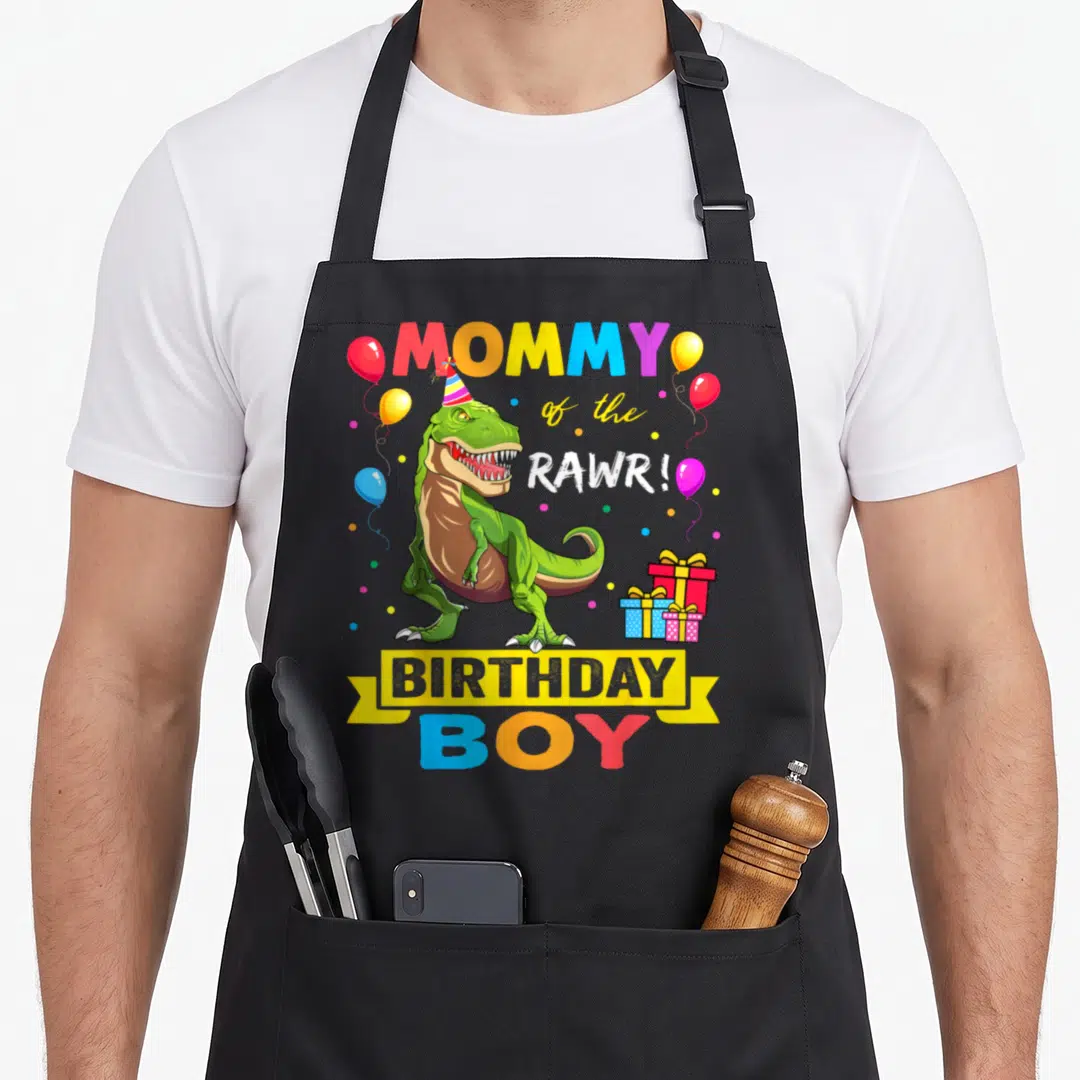 Birthday raptor dinosaur apron - Green T-Rex wearing party hat with MOMMY of the RAWR! BIRTHDAY BOY text and balloons — shown on a black cotton canvas apron against a white background
