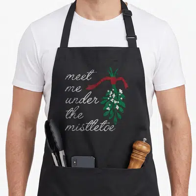 Meet christmas apron - Distressed white script reading meet me under the mistletoe next to a green sprig tied — shown on a black cotton canvas apron against a white background