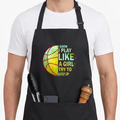 Vintage basketball boys apron - Split basketball graphic with I KNOW I PLAY LIKE A GIRL TRY TO KEEP UP text in green — shown on a black cotton canvas apron against a white background