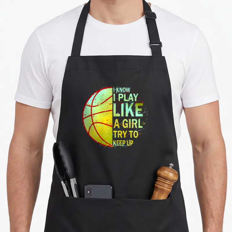 Vintage basketball boys apron - Split basketball graphic with I KNOW I PLAY LIKE A GIRL TRY TO KEEP UP text in green — shown on a black cotton canvas apron against a white background