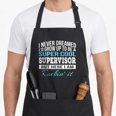 Cute supervisor apron - Stacked white and blue text reading I NEVER DREAMED SUPER COOL SUPERVISOR BUT HERE I AM Rockin — shown on a black cotton canvas apron against a white background